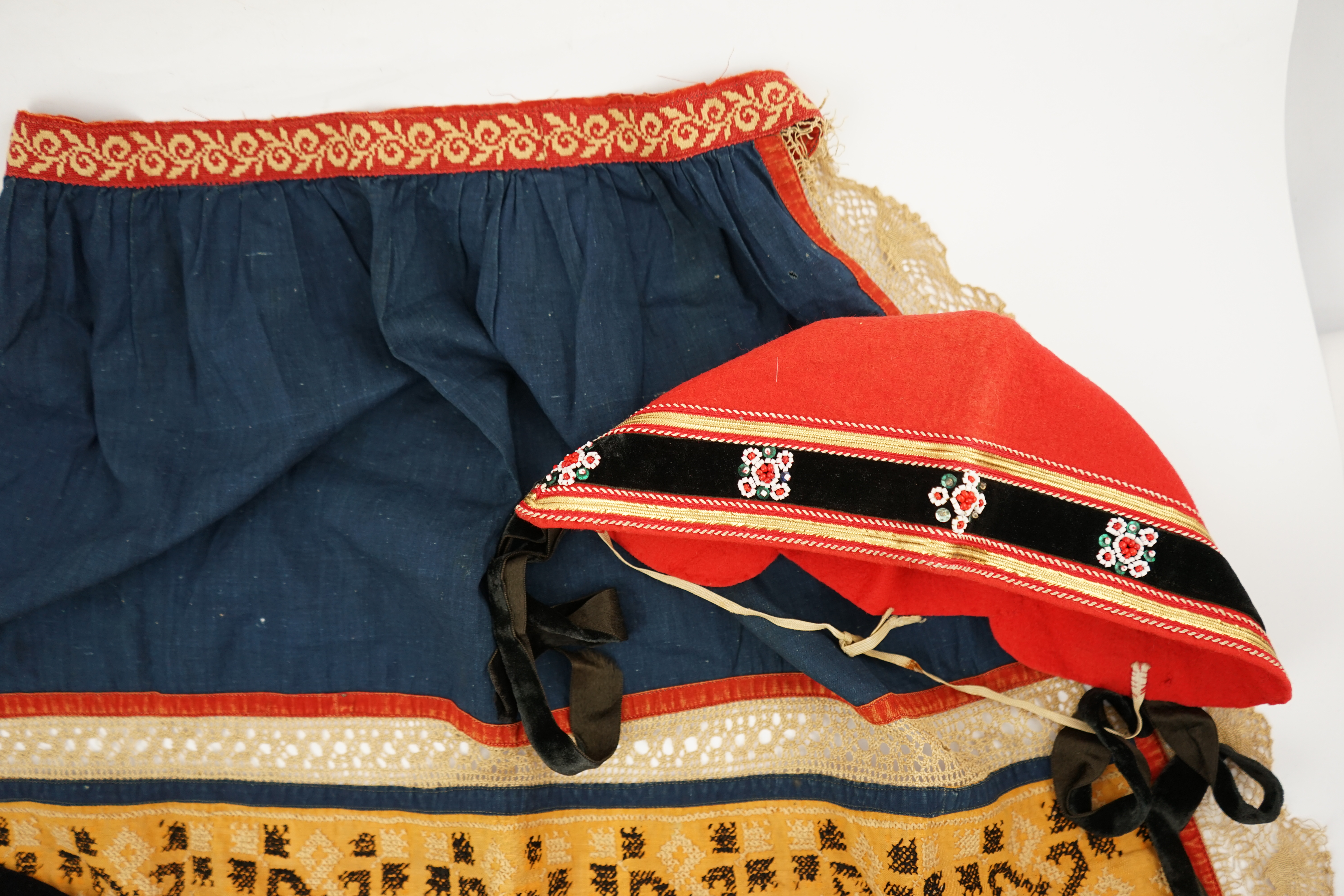 An Eastern European, possibly Ukrainian, apron and a Norwegian beaded bodice front, with child’s cap, the apron worked in rows of coloured cotton, embroidered with cross stitch patterns and lace insertions, the bodice, b